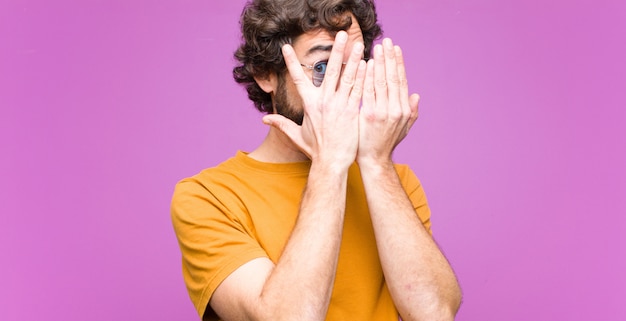 Jeune homme cool fou se sentant effrayé ou embarrassé, furtivement ou espionnant avec les yeux à moitié couverts de mains contre le mur plat