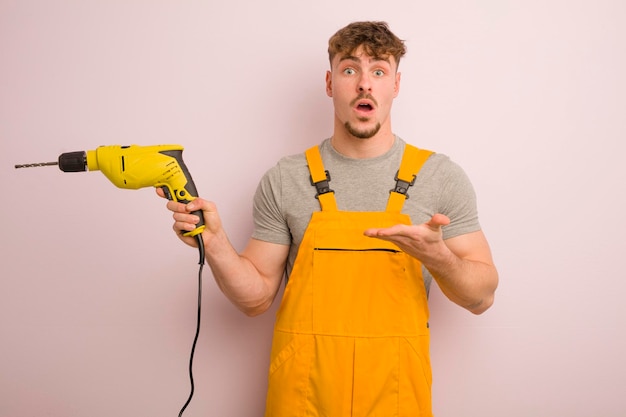 Jeune homme cool qui a l'air surpris et choqué avec la mâchoire tombée tenant un concept de bricoleur et de perceuse d'objet