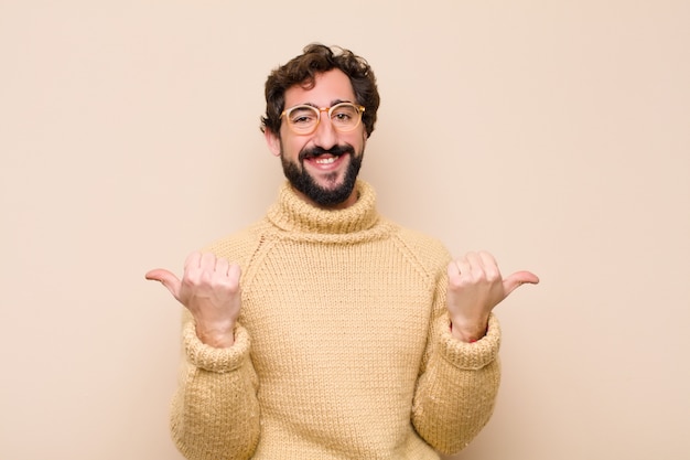 Jeune homme cool souriant joyeusement et l'air heureux, se sentant insouciant et positif avec les deux pouces contre un mur plat