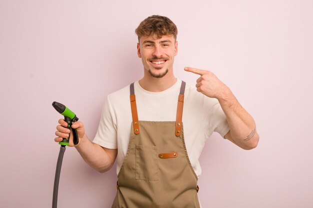 Jeune homme cool souriant en toute confiance pointant vers son propre concept de jardinier et de tuyau au large sourire