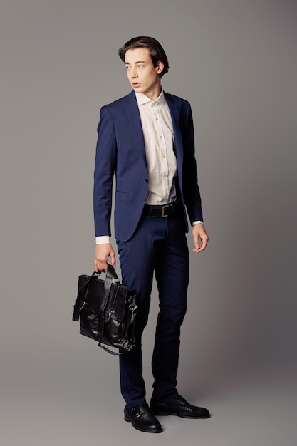 Jeune homme en costume bleu marine avec sac d'affaires noir sur mur gris