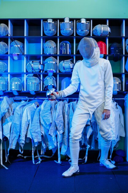 Jeune homme en costume d'escrime avec une épée à la main, sur fond bleu, néon. L'athlète s'entraîne. Sports, jeunesse, mode de vie sain.