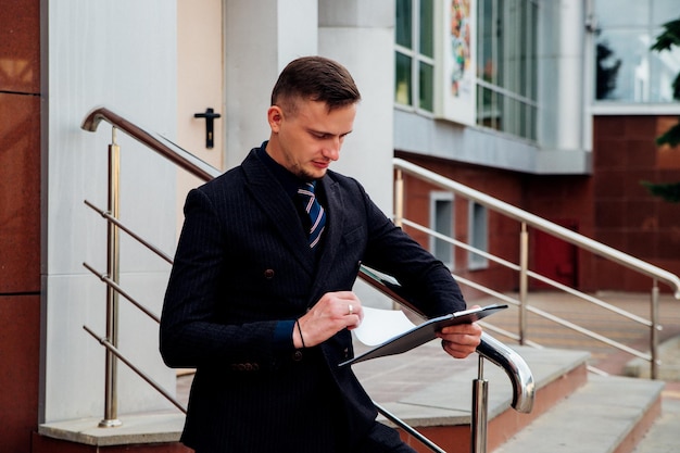 Un jeune homme en costume officiel se tient dans la rue avec des documents à la main.