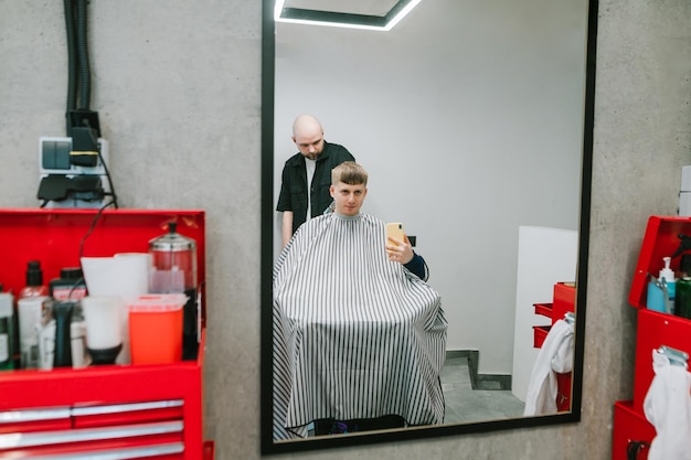 Jeune homme coupe ses cheveux dans un salon de coiffure et prend un selfie avec son homme un coiffeur dans le miroir