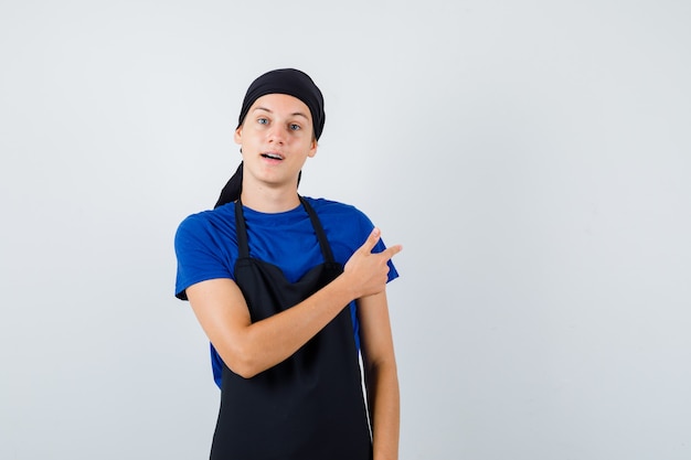 Jeune homme cuisinier pointant vers l'arrière en t-shirt, tablier et l'air perplexe, vue de face.