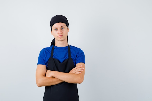 Jeune homme cuisinier en t-shirt, tablier debout avec les bras croisés et l'air confiant, vue de face.