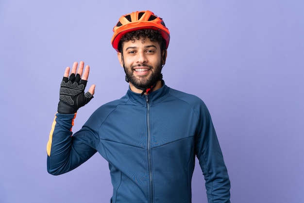 Jeune homme cycliste marocain isolé sur mur violet saluant avec la main avec une expression heureuse