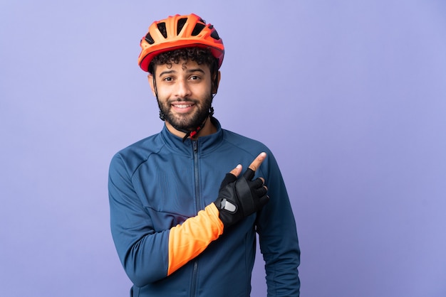 Jeune homme cycliste marocain isolé sur violet pointant vers le côté pour présenter un produit