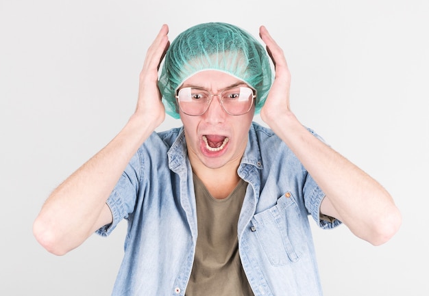 Un jeune homme dans un bonnet médical et des lunettes hurle de peur pendant l'accouchement sur un mur gris. Concept de processus d'accouchement.