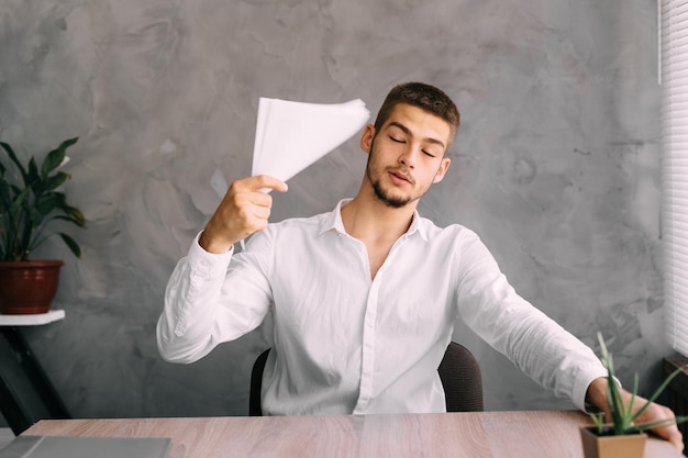 Un jeune homme dans un bureau chaud s'évente avec un morceau de papier Un gars en chemise assis à une table un travailleur fatigué se repose Chef de bureau sur le lieu de travail Concept d'entreprise