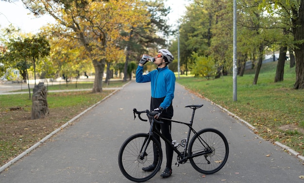 Un jeune homme dans un casque de vélo boit de l'eau d'une bouteille de fitness et tient un vélo dans ses mains. Activités sportives et livraison de nourriture dans la ville à vélo.