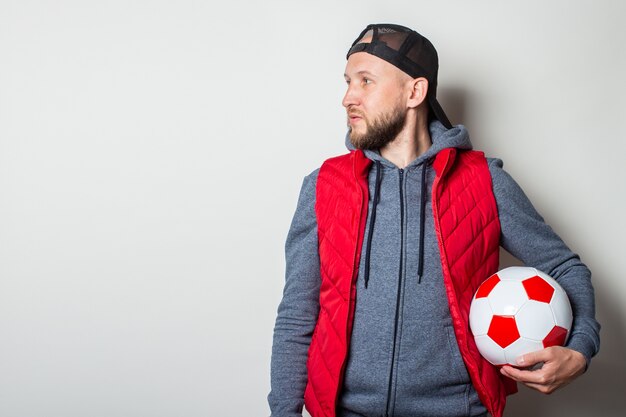 Jeune homme dans une casquette, un sweat à capuche et un gilet est titulaire d'un ballon de football et regarde sur le côté contre un mur léger