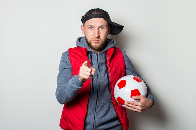 Jeune homme dans une casquette, un sweat à capuche et un gilet tient un ballon de football et pointe son doigt vers le spectateur