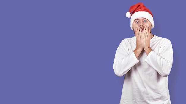 Un jeune homme dans un chapeau de père Noël couvre sa bouche avec ses mains en état de choc