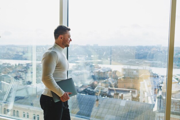 Un jeune homme dans un espace de travail moderne se tient près d'une grande fenêtre Un homme en tenue décontractée avec un ordinateur portable dans les mains Travail à distance Joli jeune manager