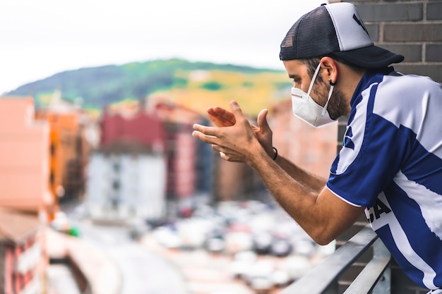 Un jeune homme dans un masque et une chemise bleue et blanche applaudissant sur le balcon à 8 heures de l'après-midi. Coronavirus pandémique en Espagne