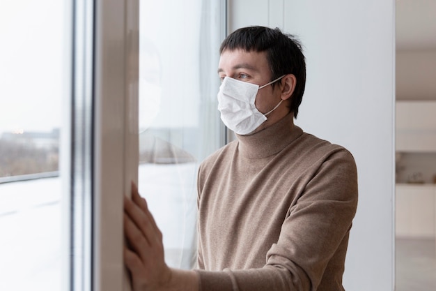 Un jeune homme dans un masque médical regarde par la fenêtre. La solitude dans l'isolement. Pandémie de Coronavirus.