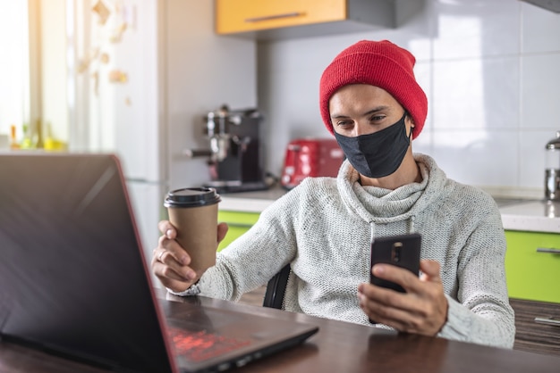 Un jeune homme dans un masque noir utilise un ordinateur portable et un téléphone tout en travaillant à la maison