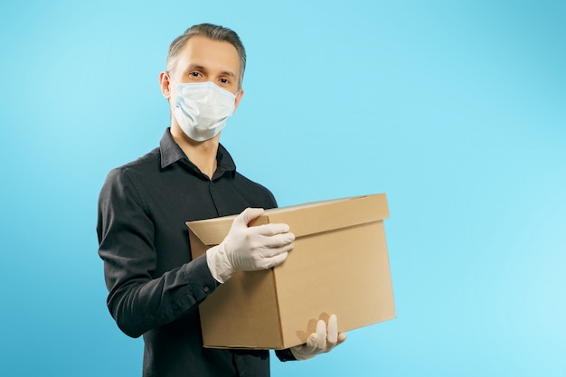 Jeune homme dans un masque de protection médical et des gants tenant une boîte sur un fond bleu. Livraison sécurisée