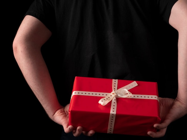 Un jeune homme dans un t-shirt noir tient un cadeau rouge derrière son dos sur fond noir