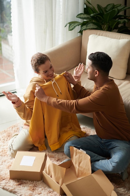 Jeune homme déballant la boîte avec un nouveau pull et le donnant à sa petite amie alors qu'ils étaient assis dans la pièce où ils faisaient des achats en ligne