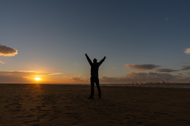 Jeune homme, debout, levant les bras, regardant le coucher de soleil sur la plage