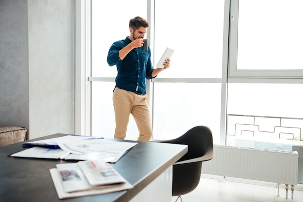 Jeune homme debout près de la fenêtre et boire du café en lisant le journal