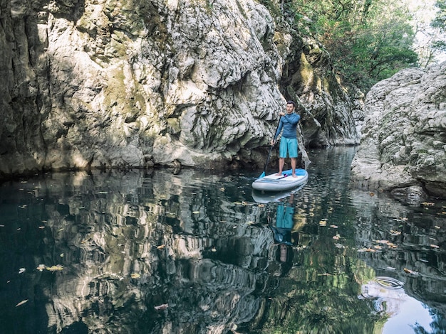 Jeune homme debout sur SUP avec pagaie et flottant sur l'eau de la rivière dans un canyon rocheux