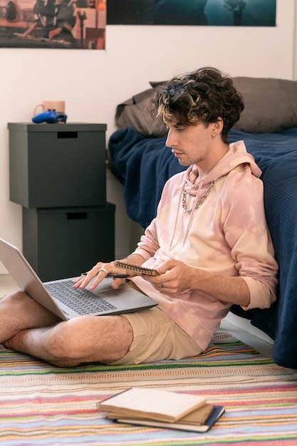 Jeune homme décontracté assis sur le sol près du lit et utilisant un ordinateur portable