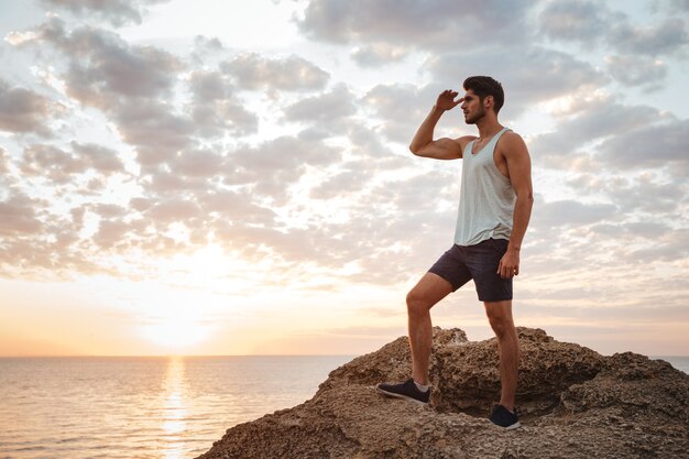 Jeune homme décontracté debout sur le rocher de la montagne au bord de la mer et regardant le coucher du soleil