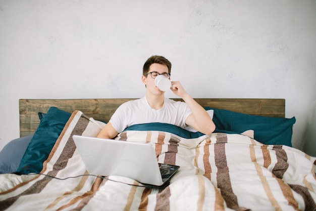 Jeune homme décontracté utilisant un ordinateur portable au lit à la maison et buvant du café.