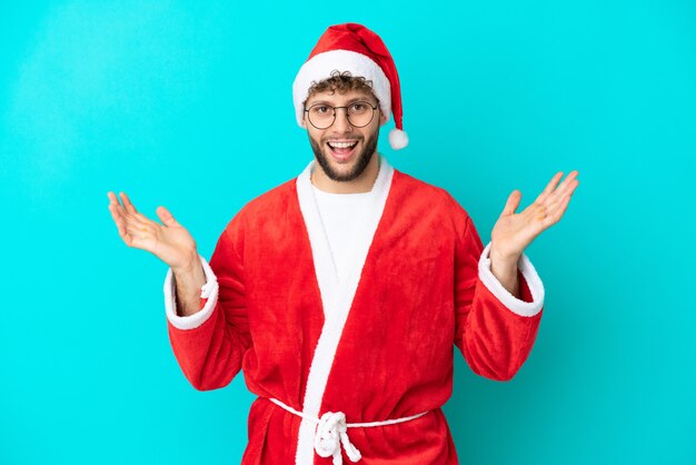 Jeune homme déguisé en Père Noël isolé sur fond bleu avec une expression faciale choquée