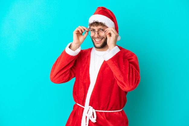 Jeune homme déguisé en Père Noël isolé sur fond bleu avec des lunettes et surpris