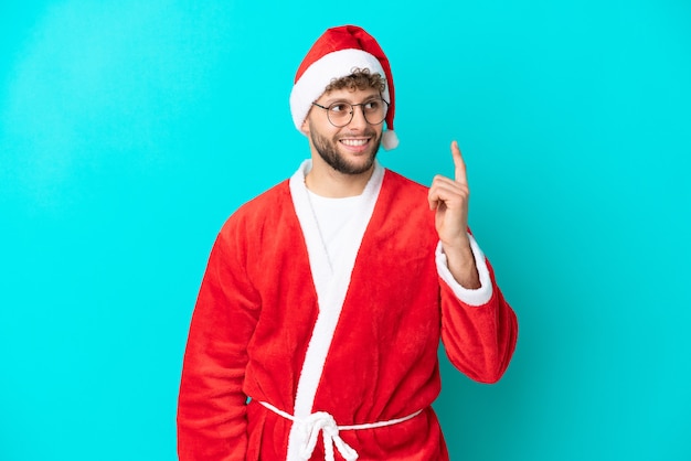 Jeune homme déguisé en Père Noël isolé sur fond bleu pensant à une idée pointant le doigt vers le haut