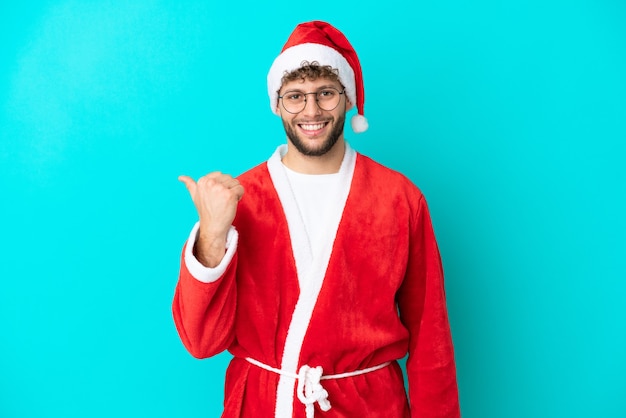 Jeune homme déguisé en Père Noël isolé sur fond bleu pointant vers le côté pour présenter un produit