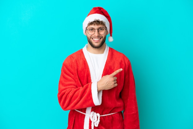 Jeune homme déguisé en Père Noël isolé sur fond bleu pointant vers le côté pour présenter un produit