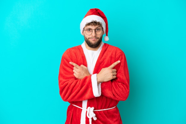 Jeune homme déguisé en Père Noël isolé sur fond bleu pointant vers les latéraux ayant des doutes