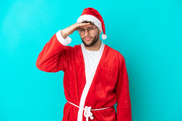 Jeune homme déguisé en Père Noël isolé sur fond bleu regardant loin avec la main pour regarder quelque chose