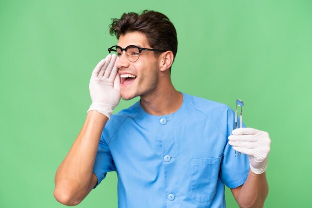 Jeune homme dentiste tenant des outils sur fond isolé criant avec la bouche grande ouverte sur le côté