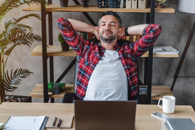 Jeune homme détendu se reposant du travail sur un ordinateur portable tenant les mains derrière la tête dans le bureau à domicile