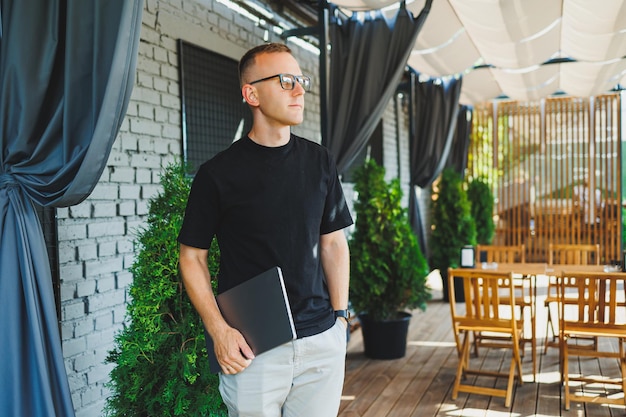 Jeune homme développeur indépendant profitant d'une pause-café sur la terrasse d'été d'un café confortable travaillant à distance sur un ordinateur portable souriant regardant un ordinateur portable