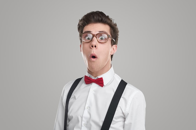 Jeune homme diligent en chemise blanche avec nœud papillon et bretelles regardant la caméra à travers des lunettes en état de choc sur fond gris