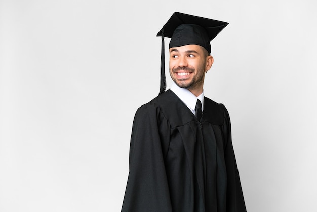 Jeune homme diplômé universitaire sur fond blanc isolé avec les bras croisés et heureux