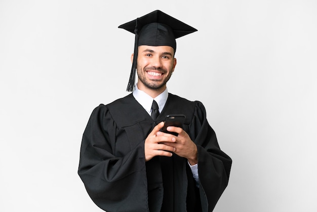 Jeune homme diplômé universitaire sur fond blanc isolé envoyant un message avec le mobile