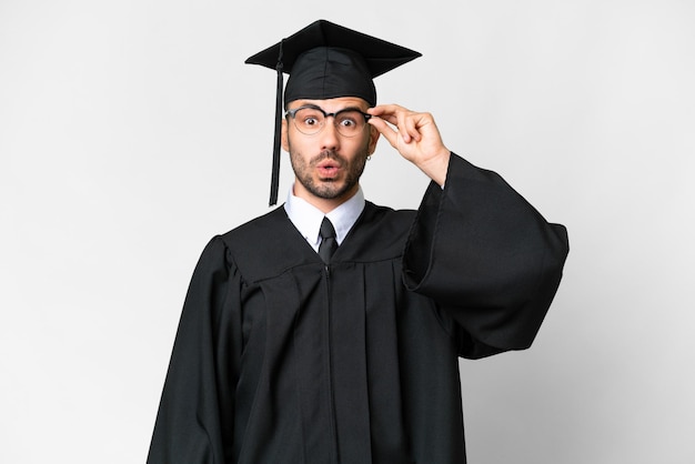 Jeune homme diplômé universitaire sur fond blanc isolé avec des lunettes et surpris