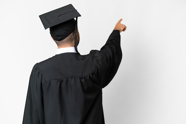 Jeune homme diplômé universitaire sur fond blanc isolé pointant vers l'arrière avec l'index