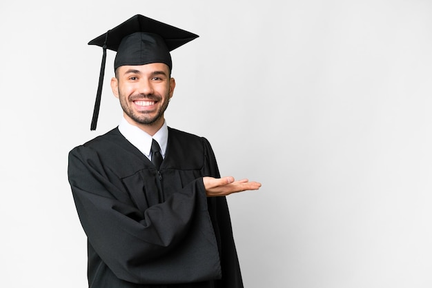 Jeune homme diplômé universitaire sur fond blanc isolé présentant une idée tout en regardant en souriant vers