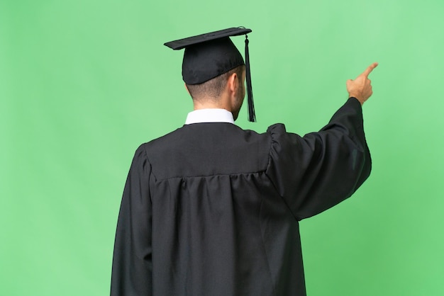 Jeune homme diplômé universitaire sur fond isolé pointant vers l'arrière avec l'index