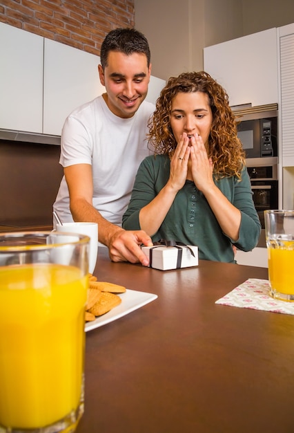 Jeune homme donnant une boîte-cadeau à sa petite amie surprise tout en prenant son petit-déjeuner dans la cuisine de la maison