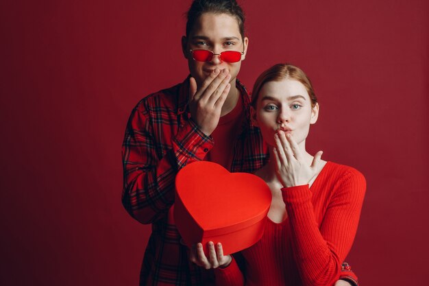 Jeune homme donne à sa petite amie une boîte en forme de coeur à la Saint-Valentin sur fond rouge studio